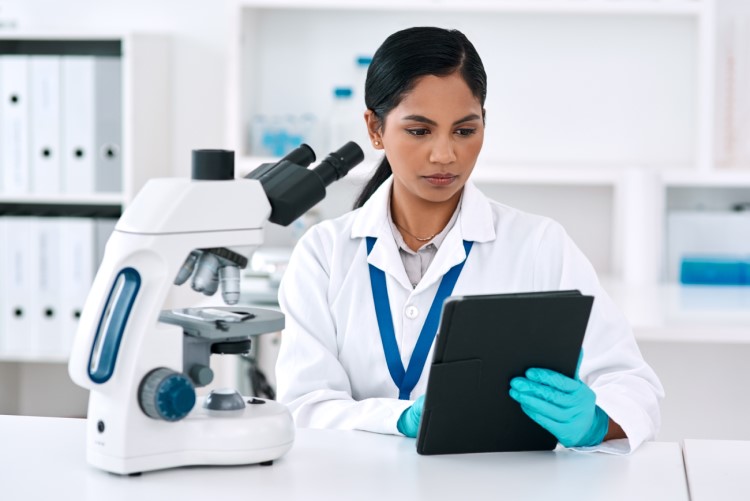 Photo depicts: a female scientist sits at a bench with a microscope, reviewing information on a tablet